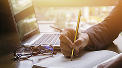 An open laptop on a desk, a person writes in a notebook next to it.