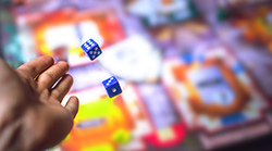 A hand tosses two blue dice over an out-of-focus game board.